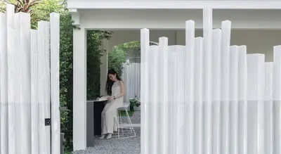 a woman is sitting at a table in front of a white fence at InJoy Snow Hotel in Ban Chuat Plai Mai